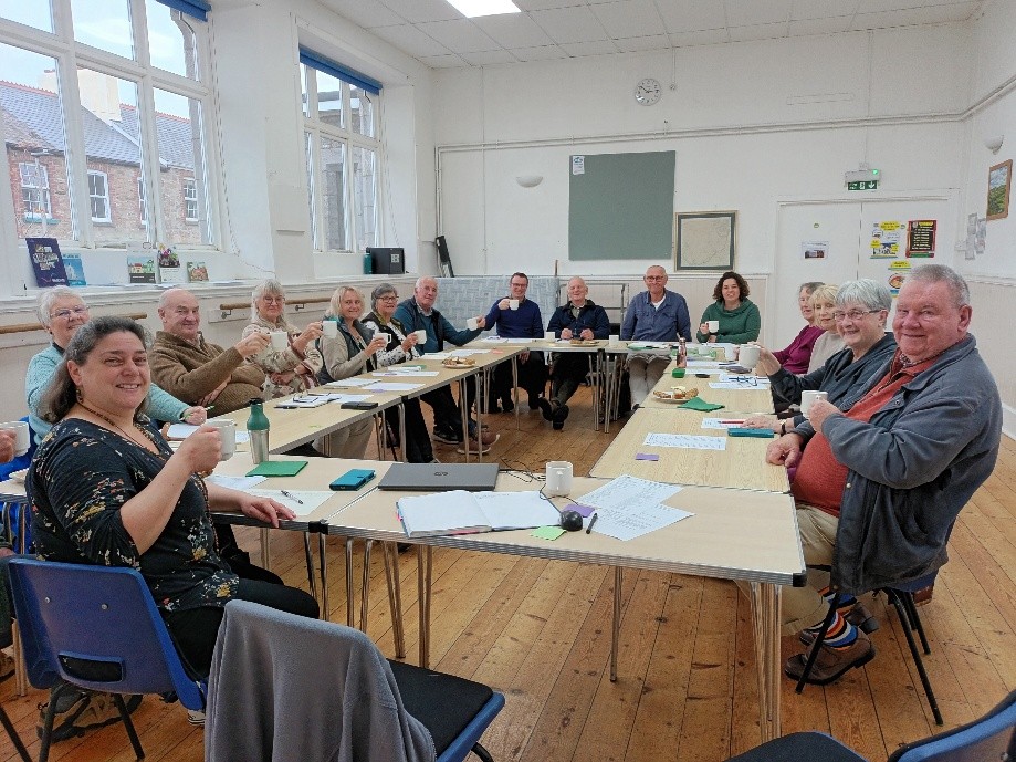 Photo of lots of people sat around a table in a village hall - at a cluster meeting