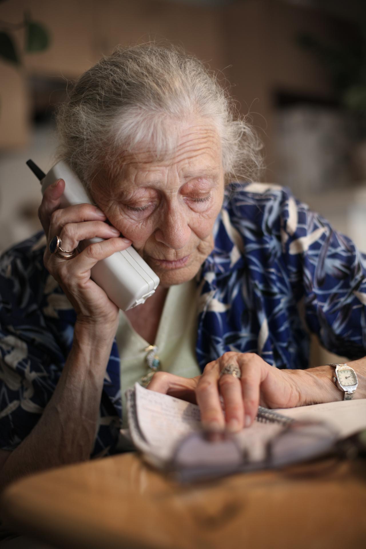 Elderly lady holding a cordless phone in her hand to her ear, and looking at some information in a book on a table
