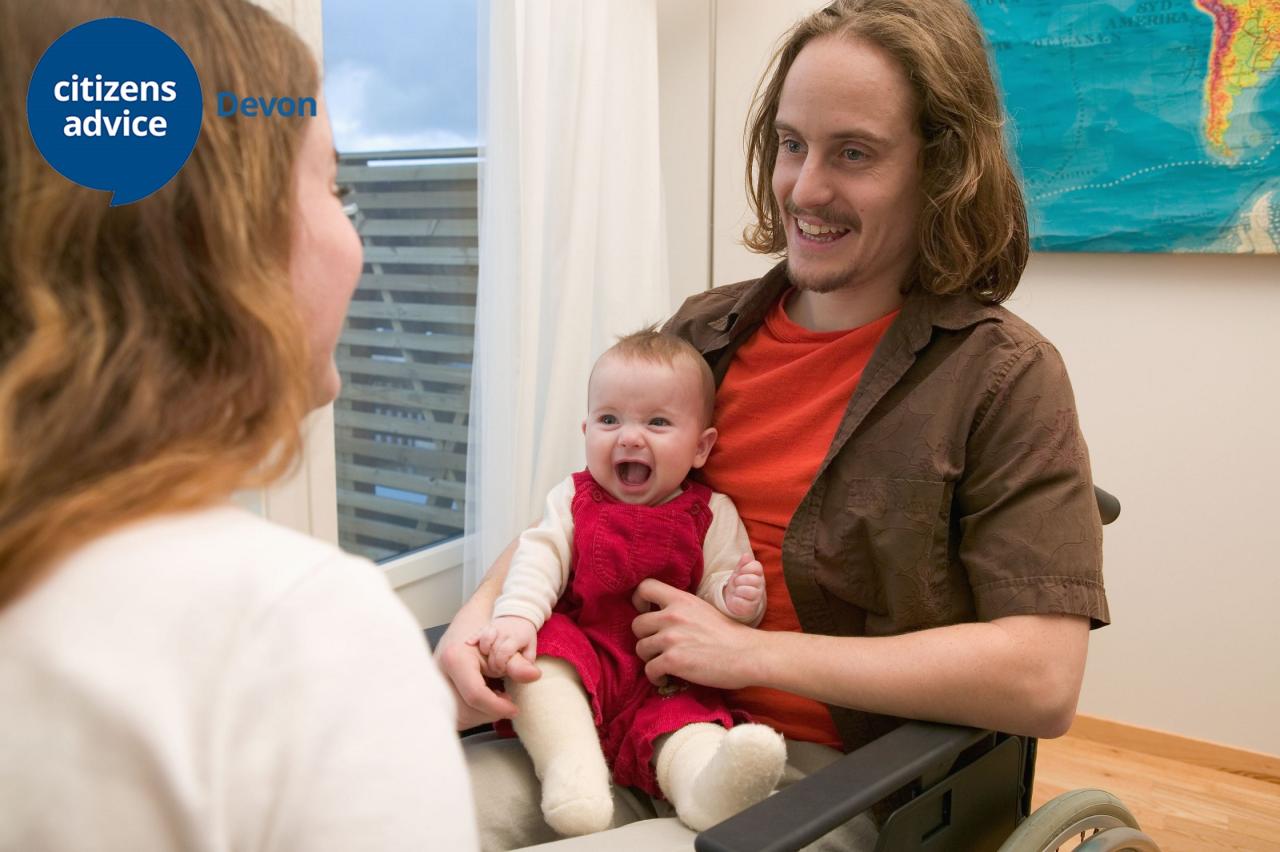 Young white male in a wheelchair smiling and talking to a lady, holding a young baby on his lap