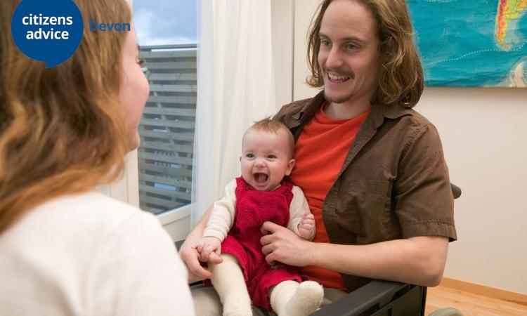 Young white male in a wheelchair smiling and talking to a lady, holding a young baby on his lap