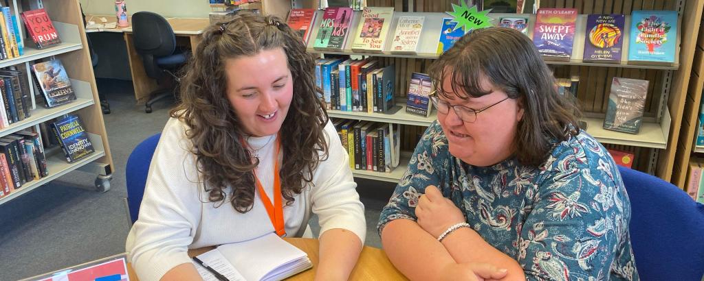 Two women sat at a table in a library looking at a tablet device