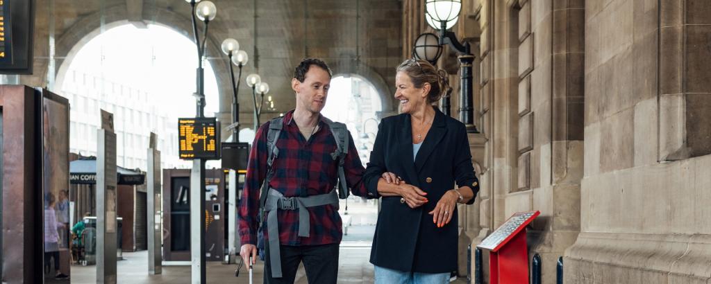 Man with white walking aid, visually impaired, with woman, walking through a train station